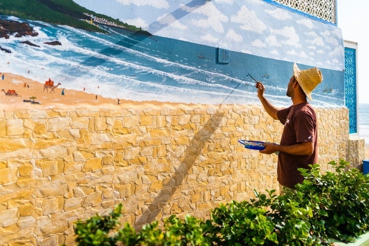 A man painting a mural on a wall. The mural is of a seascape and the man is wearing a straw hat