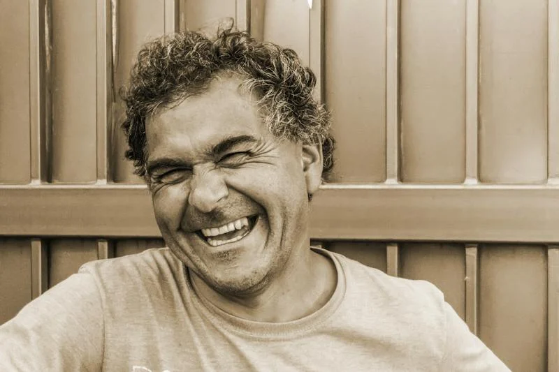 curly haired guy in sepia tone standing against a fence laughing