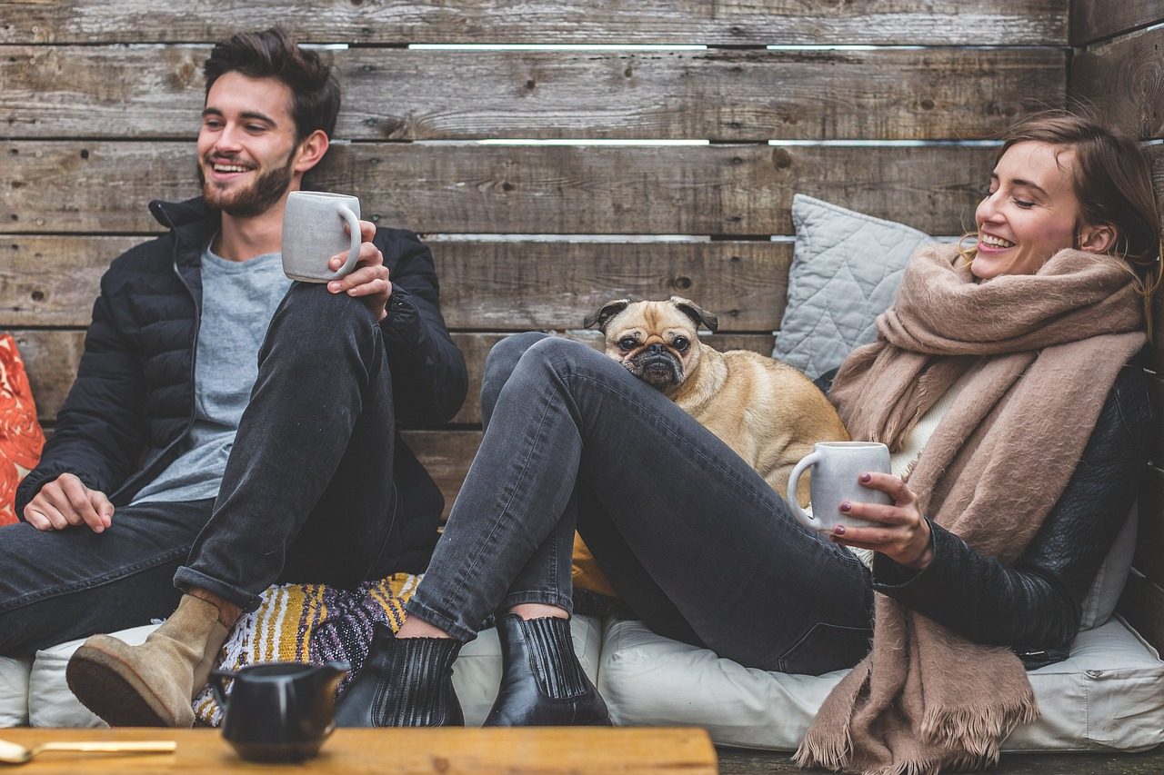 A couple laughing in a wooden cabin sharing a cup of coffee with their dog