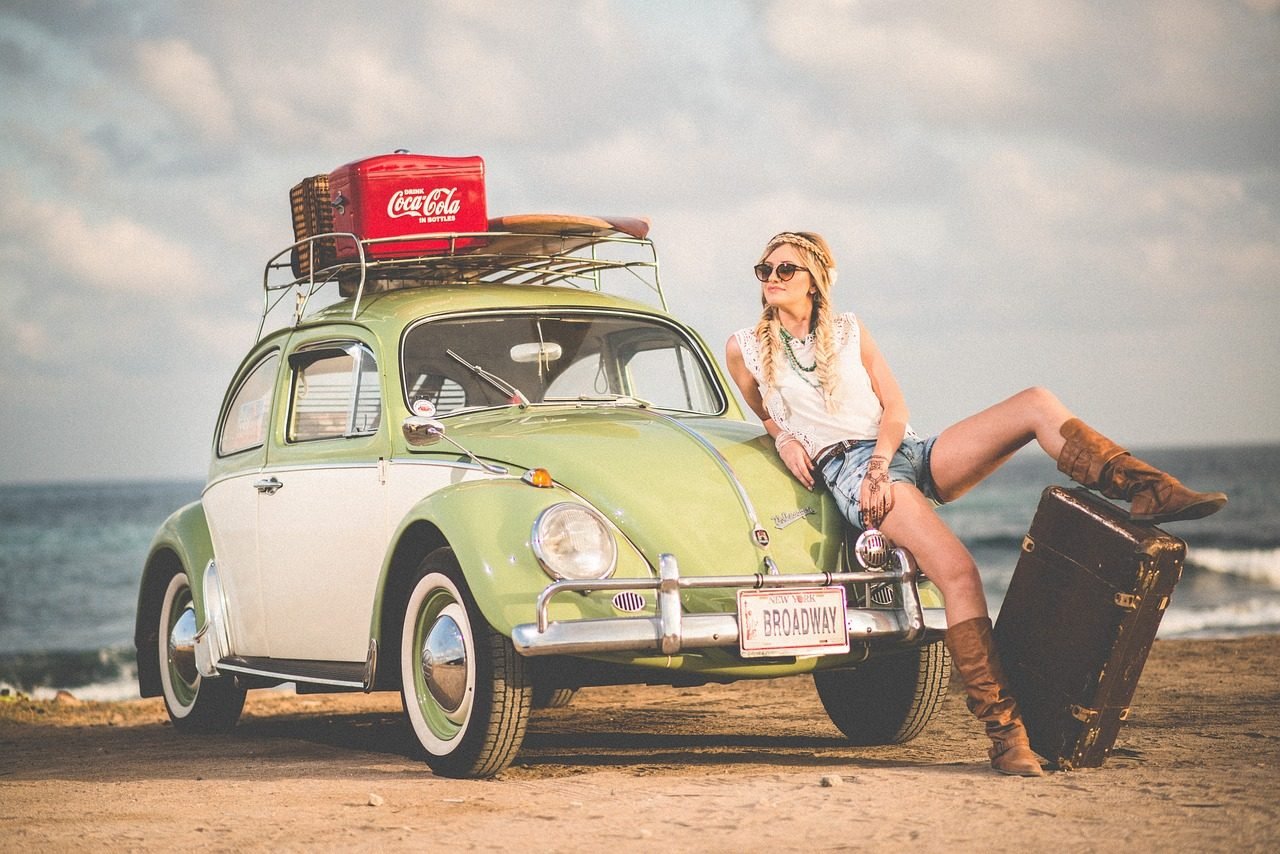 A fashion model leaning on a green BMW Beetle and resting her leg on a suitcase at the beach