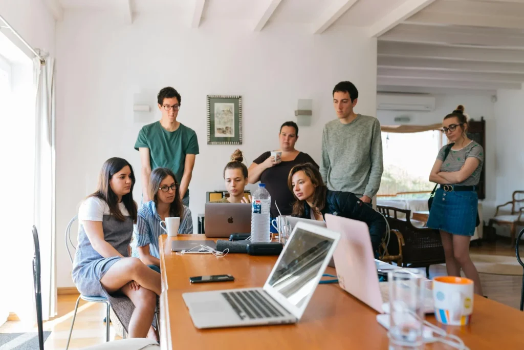 A class of people staying around as a girl demonstrates something on her laptop
