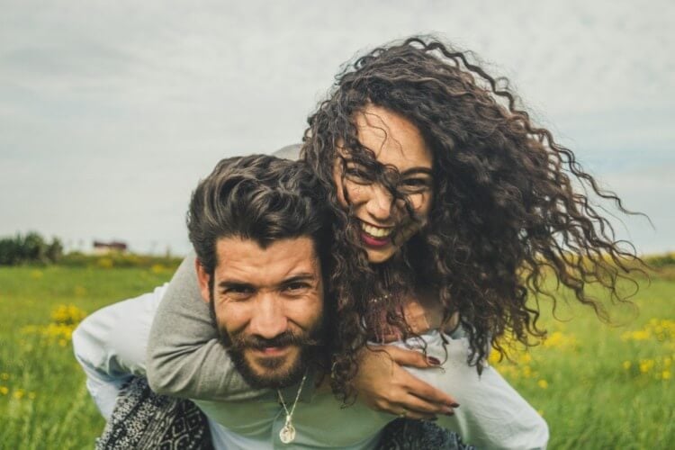 smiling couple photo of woman piggybacking on man's back. both stand in grassy field.